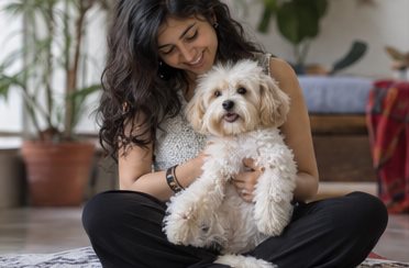A woman sits indoors and holds a small white dog.