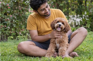A man seated on grass beside a small brown dog in a garden