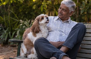 An older man sits in a garden chair as a small brown-and-white dog rests beside him