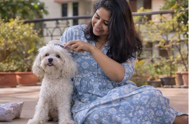 A woman sits beside a small dog in a leafy outdoor setting.
