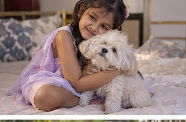 A girl sits on a bed and holds a small fluffy dog close.