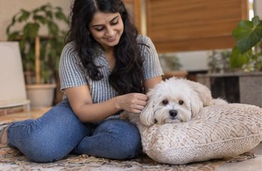 A woman sits on the floor beside a small white dog on a cushion.